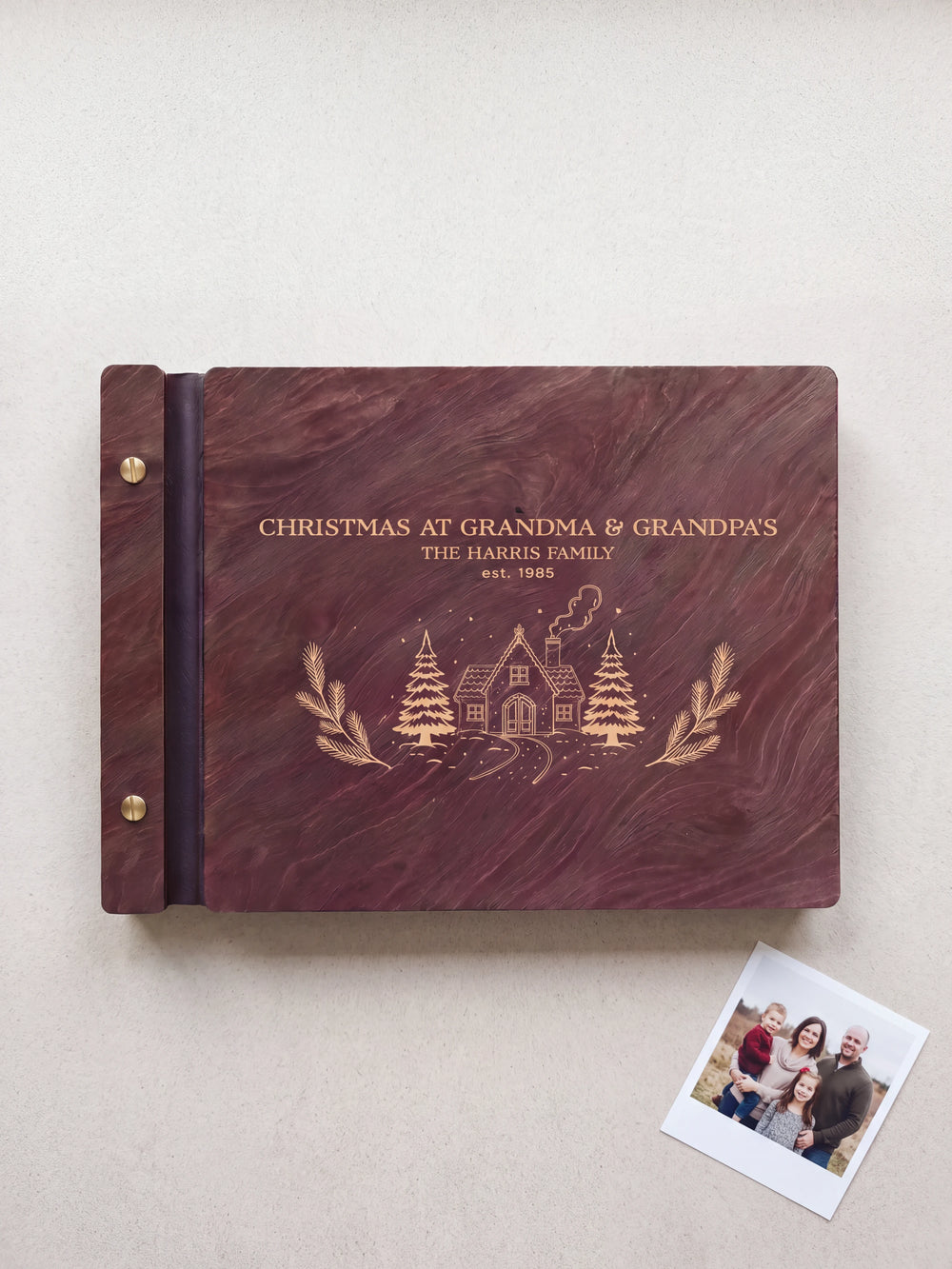 Dark Jacobean wooden album on pale fabric, with cream engraving of a snowy cottage and the words Christmas at Grandma & Grandpa's, The Harris Family, est. 1985; two gold-tone screws line the left edge