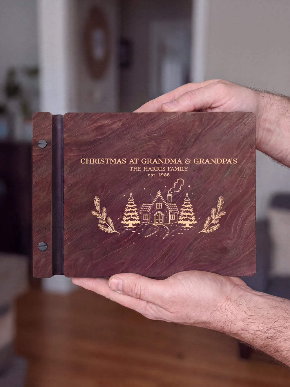 Two hands hold a Jacobean album facing the camera, the cream Christmas cottage engraving filling the cover with a personalized heading, family name and year