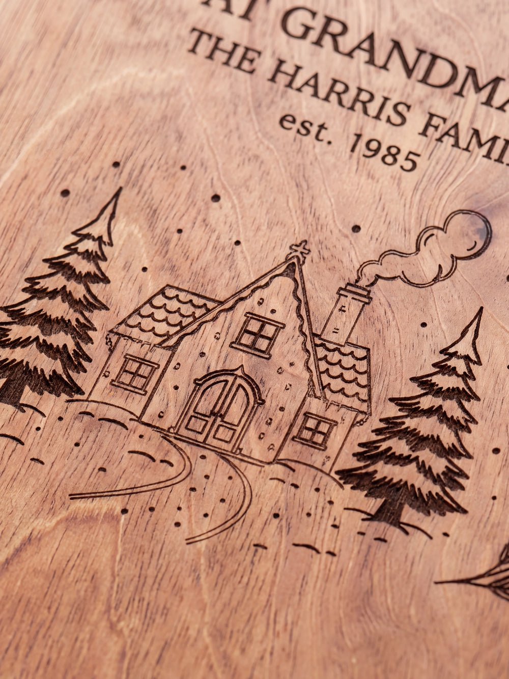 Close-up of dark brown laser engraving on Hazelnut wood, showing the snowy cottage roof, evergreen trees, pine sprigs and personalized family text against the wood grain