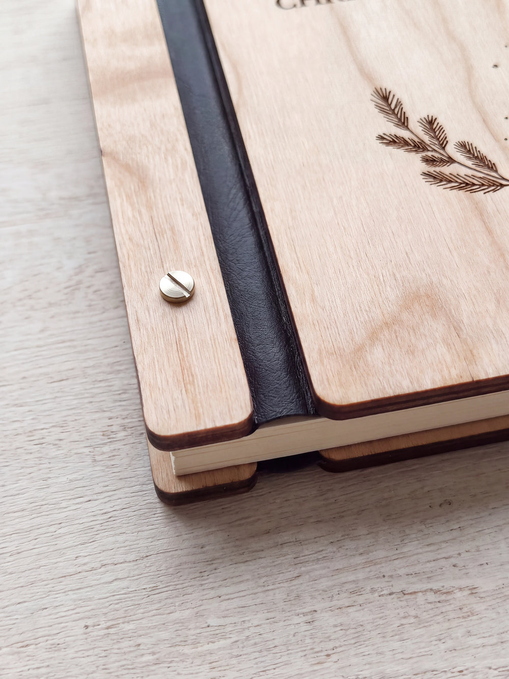 Close-up of a polished silver slotted screw in a Natural wood strip beside the black flexible binding, with the rounded cover edge and cream sheet stack below