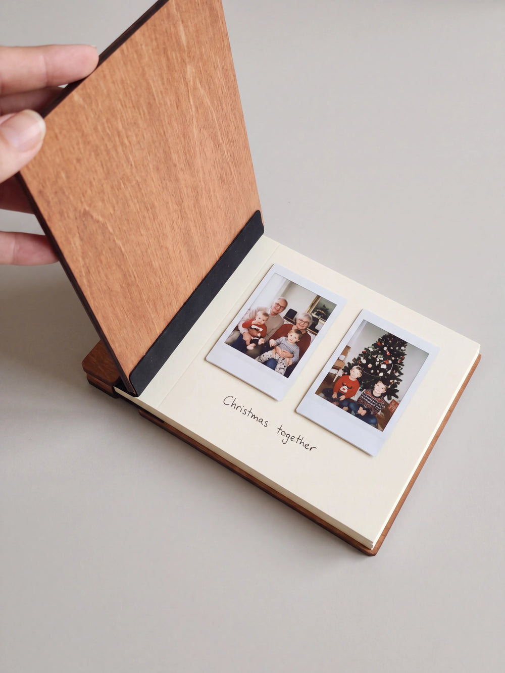 A hand holds the wooden cover open on a pale gray surface; two family instant photos and a small handwritten Christmas note sit on the cream page