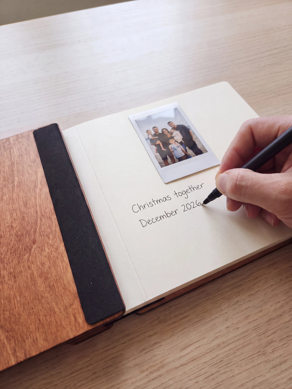 A hand writes a fine black-pen note beneath a family instant photo on a cream album page, the open wooden cover and black binding beside it