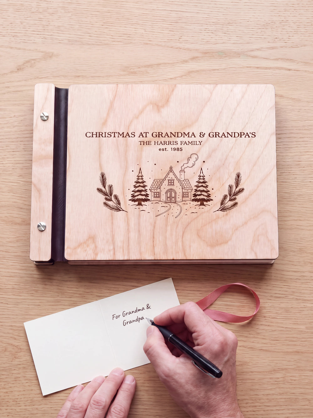 A hand writes a gift card beside a Natural wood Christmas album on a light wood table; the engraved cottage cover, silver screws and a loose ribbon are visible