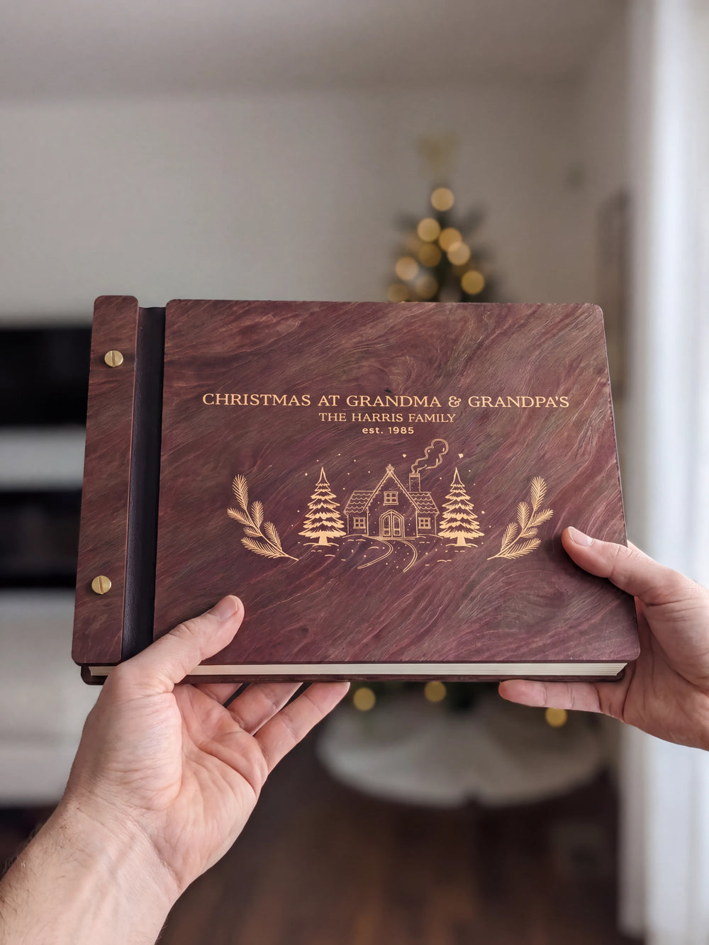 Two hands present a closed Jacobean wooden album with cream Christmas cottage engraving and gold-tone screws, a softly blurred Christmas tree behind