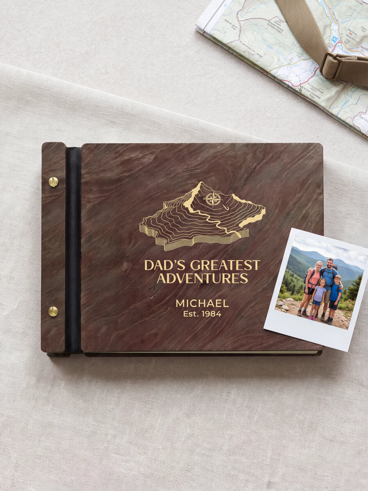 Jacobean wood Dad's Greatest Adventures photo book lying flat on a pale stone tabletop, the cover engraved with “DAD'S GREATEST ADVENTURES”, “MICHAEL” and “Est. 1984”; two gold-tone screws along the black vegan leather spine