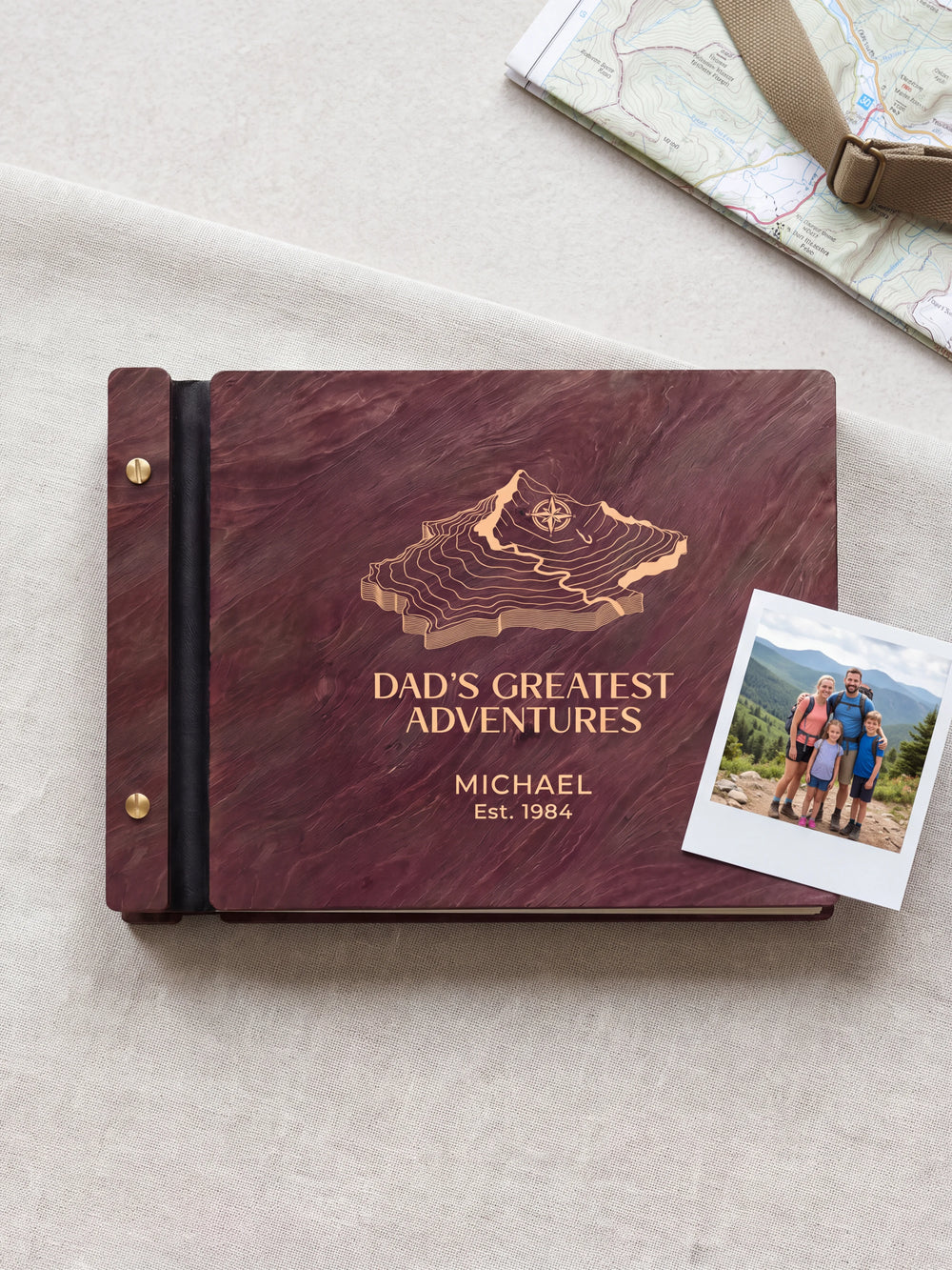 Jacobean wood Dad's Greatest Adventures photo book lying flat on a pale stone tabletop, the cover engraved with “DAD'S GREATEST ADVENTURES”, “MICHAEL” and “Est. 1984”; two gold-tone screws along the black vegan leather spine