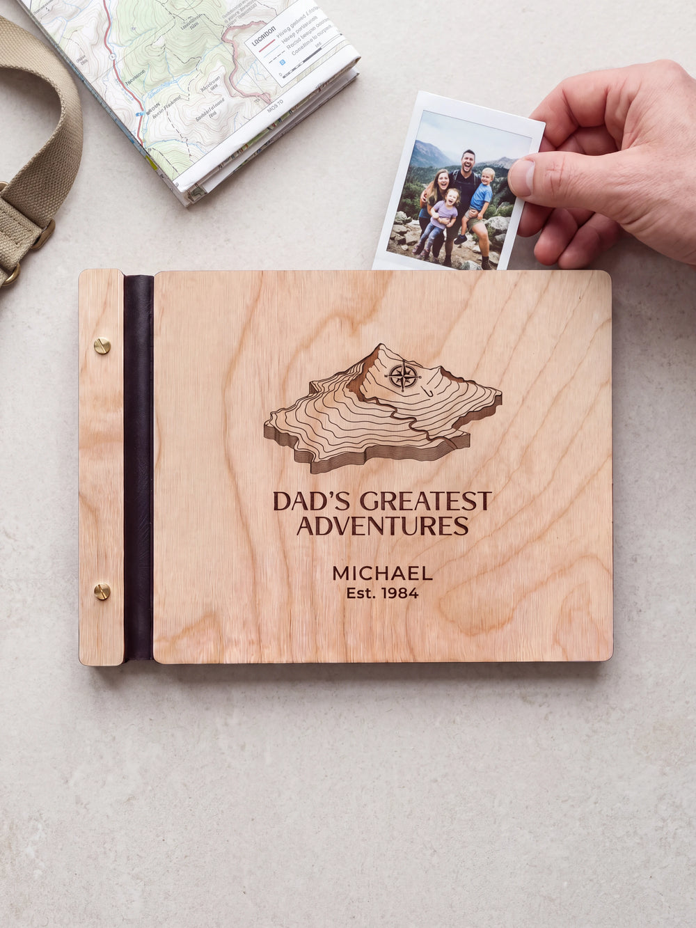 Natural wood Dad's Greatest Adventures photo book on a light table while a hand picks an instant photo to add; the engraved cover reads “DAD'S GREATEST ADVENTURES”, “MICHAEL” and “Est. 1984”