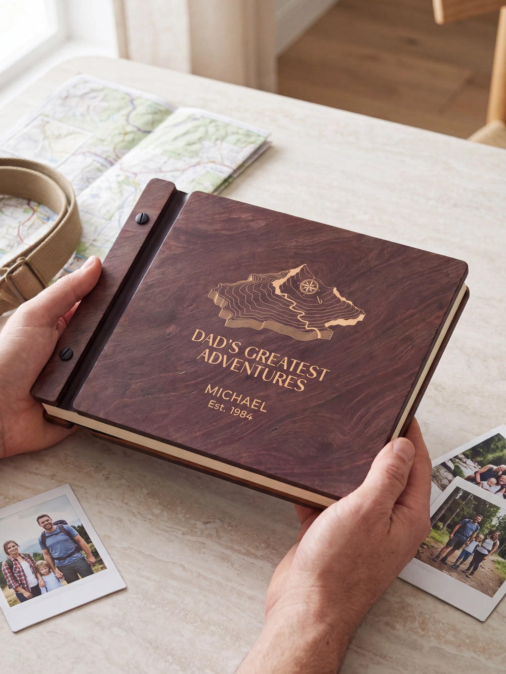 Two hands holding the Jacobean Dad's Greatest Adventures photo book up to the camera, the cream engraving of a topographic mountain map with a compass and “DAD'S GREATEST ADVENTURES”, “MICHAEL” and “Est. 1984” filling the cover