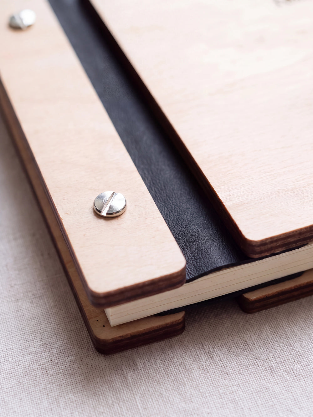 Close-up of a polished silver screw in the Natural wood strip beside the black vegan leather binding, the cream sheet stack visible below