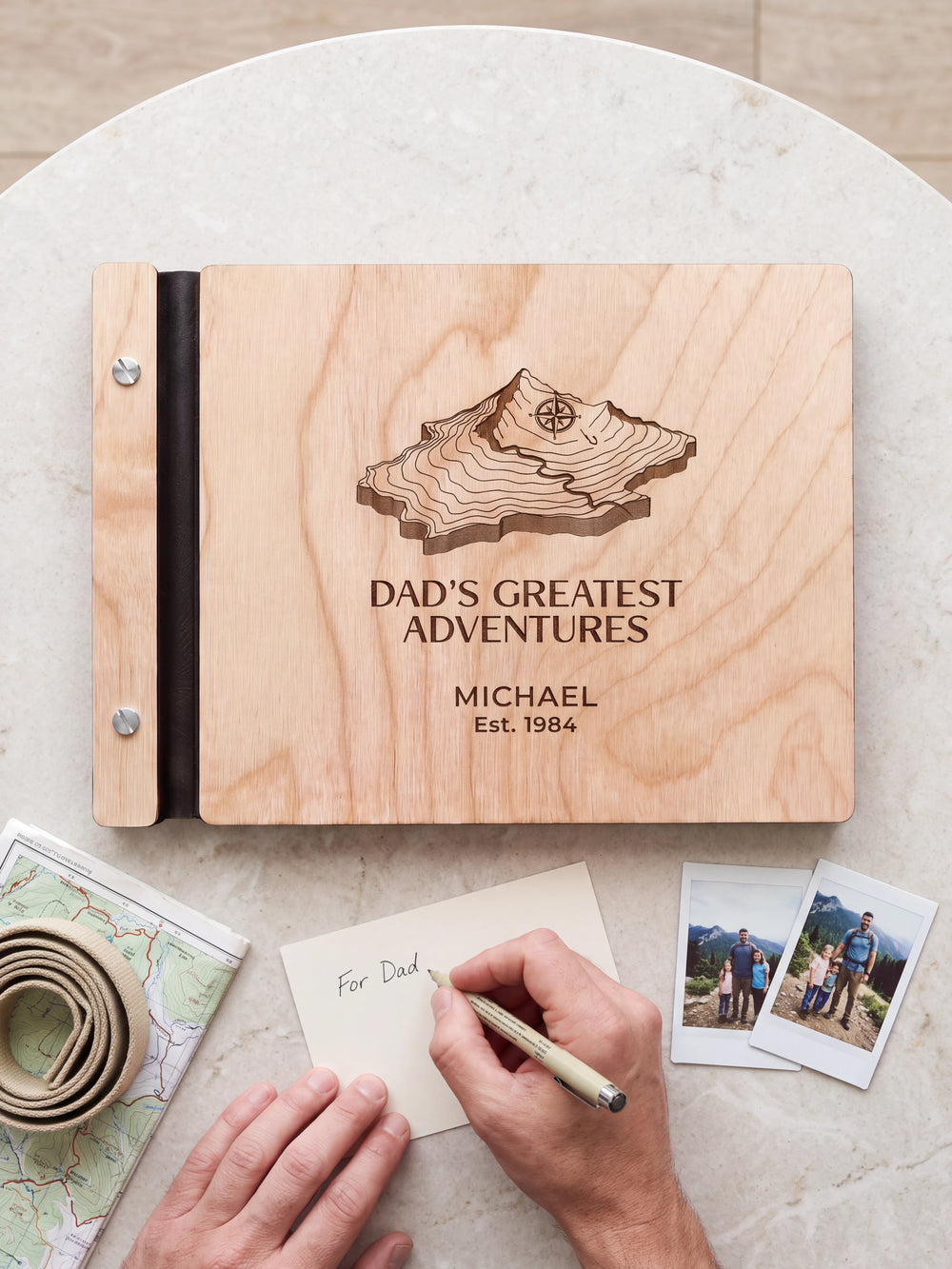 A hand writing a small gift card beside the closed Natural wood Dad's Greatest Adventures photo book; the engraved cover and silver screws are visible