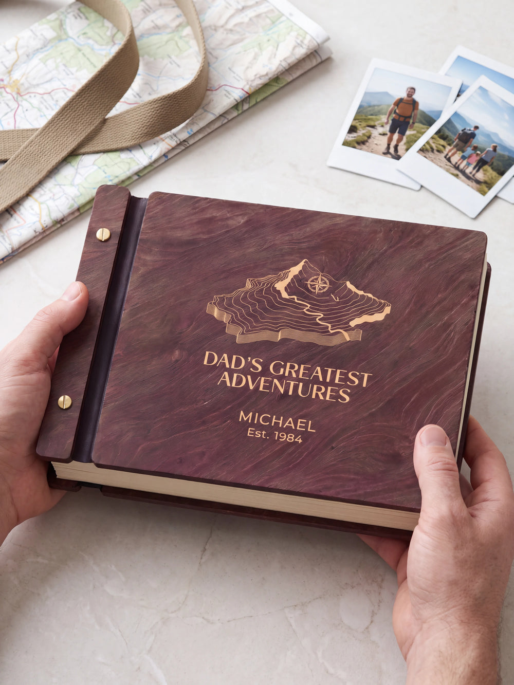 Two hands presenting the closed Jacobean Dad's Greatest Adventures photo book with its cream engraving of a topographic mountain map with a compass, a folded hiking map and canvas strap beside them