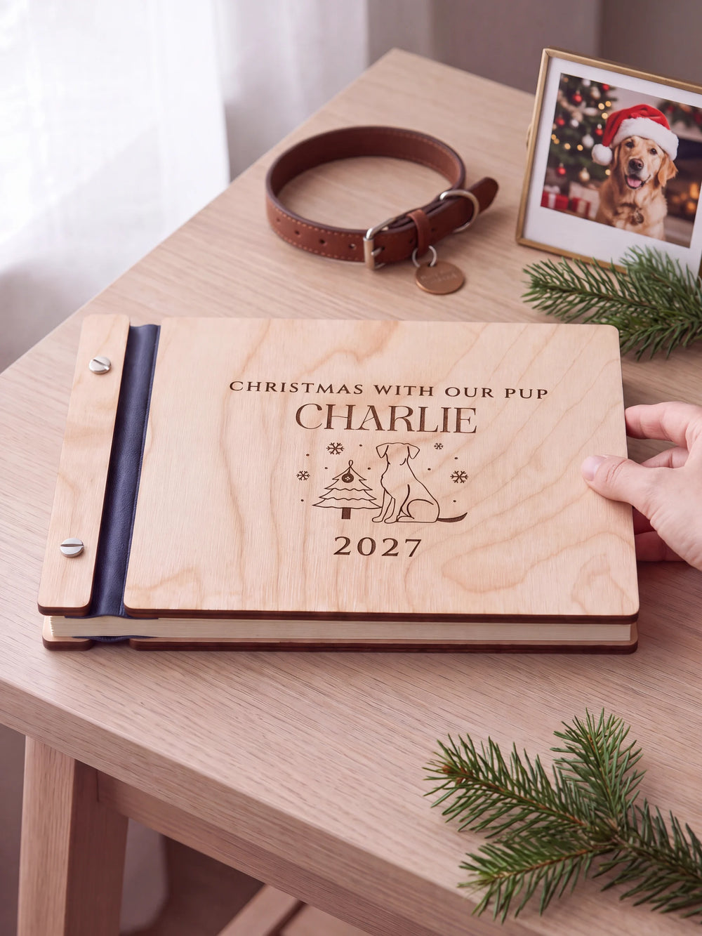 Natural wooden album with brown laser engraving of dog beside Christmas tree and snowflakes, shown in a close lifestyle view with navy blue binding and silver screws. The cover reads CHRISTMAS WITH OUR PUP.