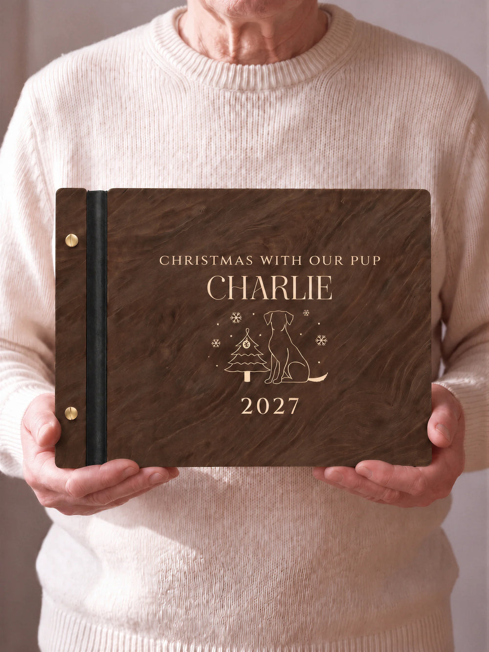 Two hands hold a Jacobean wooden photo album with the CHRISTMAS WITH OUR PUP cover facing the camera. Cream engraved artwork, the narrow binding and gold screws are visible.