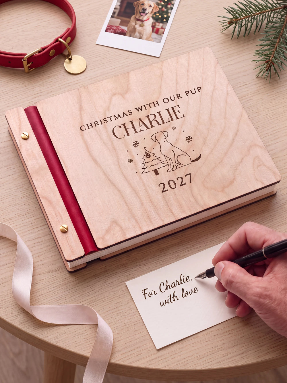 A Natural wooden album with the CHRISTMAS WITH OUR PUP engraving, cherry red binding and gold screws. A hand writes on a separate gift card beside the book.