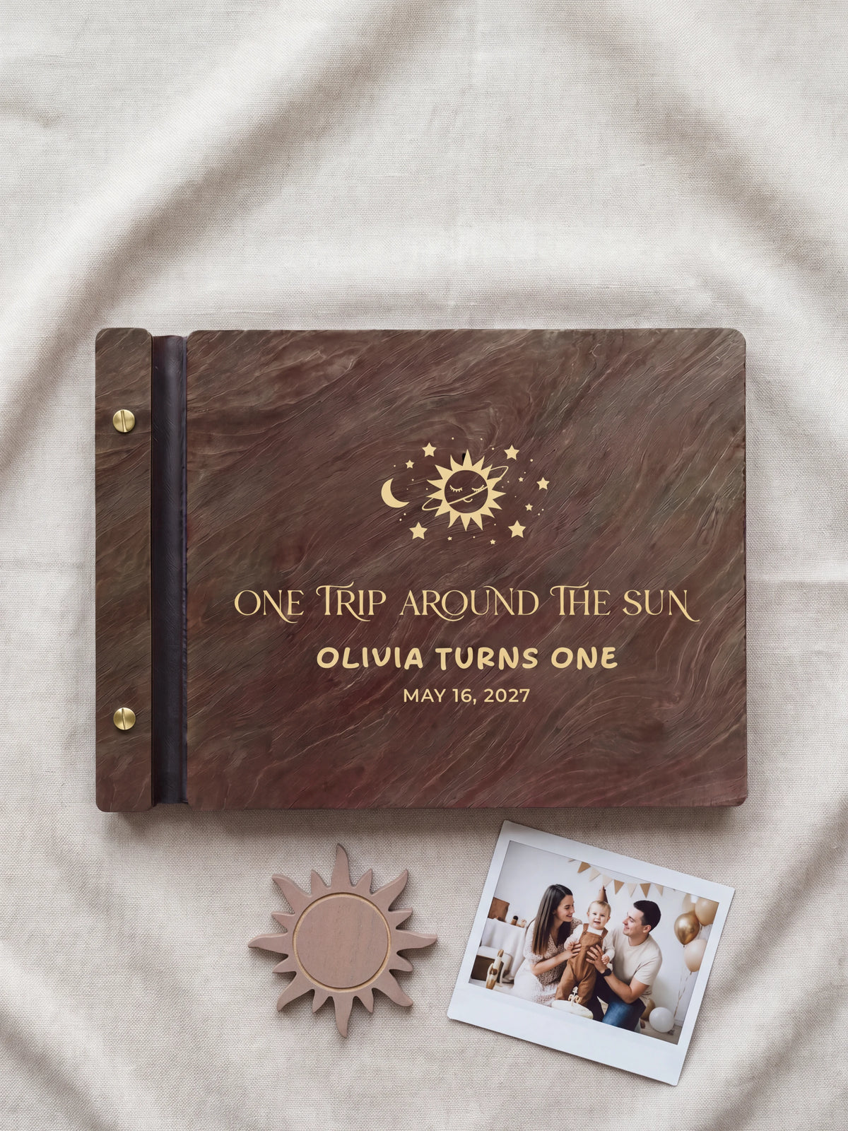 Jacobean wood first birthday photo guest book lying flat on ivory fabric, the cover engraved with “ONE TRIP AROUND THE SUN”, “OLIVIA TURNS ONE” and “MAY 16, 2027”; two gold-tone screws along the black vegan leather spine