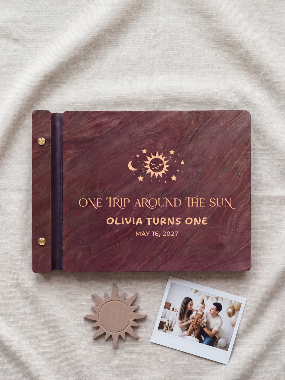 Jacobean wood first birthday photo guest book lying flat on ivory fabric, the cover engraved with “ONE TRIP AROUND THE SUN”, “OLIVIA TURNS ONE” and “MAY 16, 2027”; two gold-tone screws along the black vegan leather spine