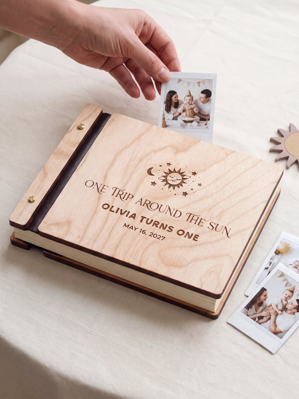 Natural wood first birthday photo guest book on a light table while a hand picks an instant photo to add; the engraved cover reads “ONE TRIP AROUND THE SUN”, “OLIVIA TURNS ONE” and “MAY 16, 2027”