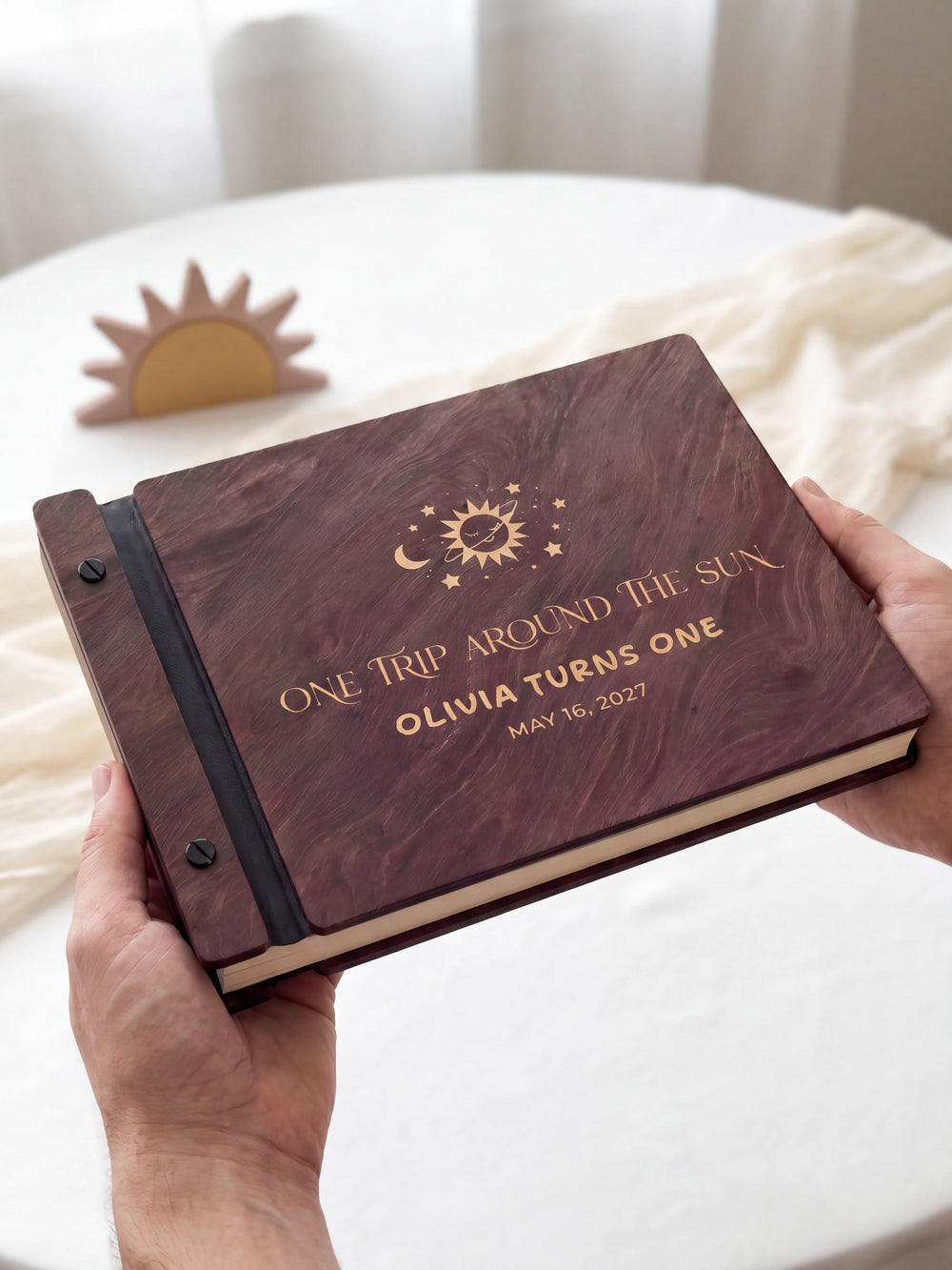 Two hands holding the Jacobean first birthday photo guest book up to the camera, the cream engraving of a smiling sun with a crescent moon and stars and “ONE TRIP AROUND THE SUN”, “OLIVIA TURNS ONE” and “MAY 16, 2027” filling the cover