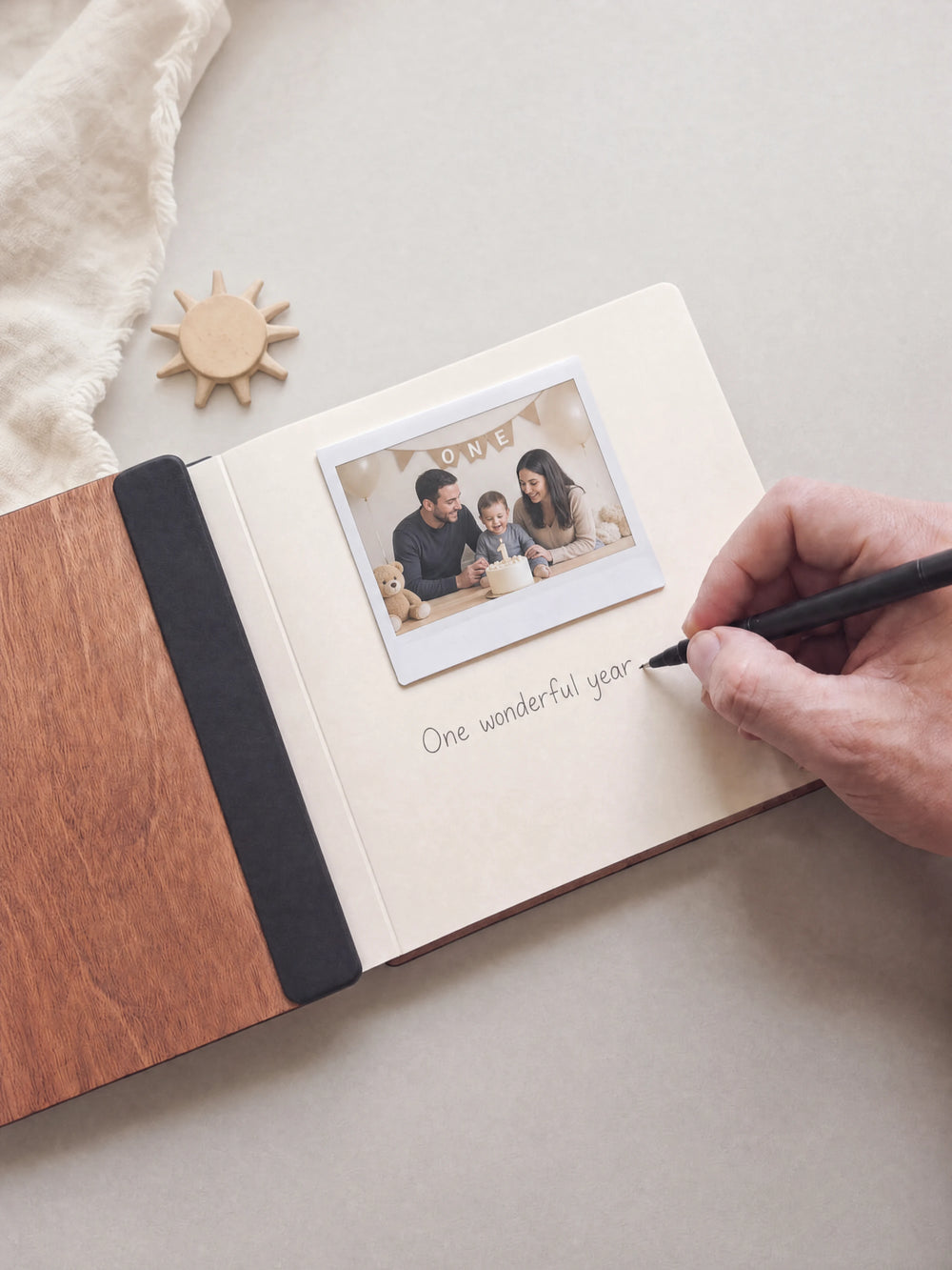 A hand writing a note in fine black pen beneath an instant photo on the cream page of the open Hazelnut first birthday photo guest book