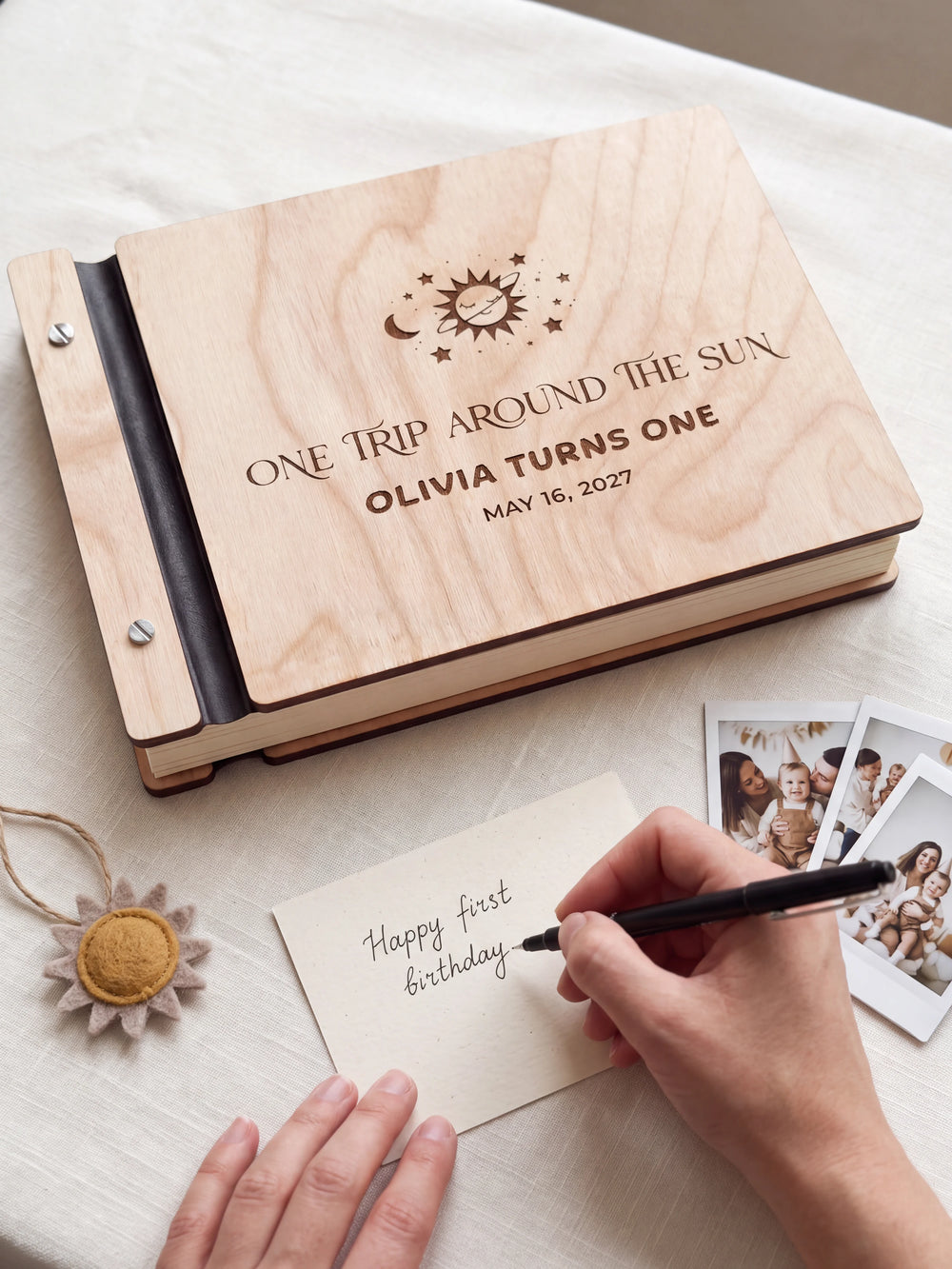A hand writing a small gift card beside the closed Natural wood first birthday photo guest book; the engraved cover and silver screws are visible