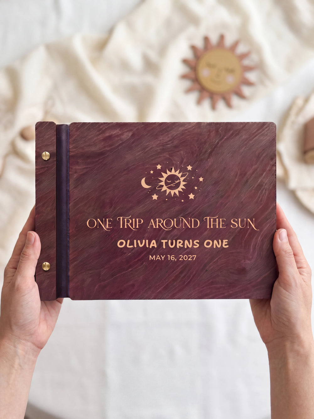 Two hands presenting the closed Jacobean first birthday photo guest book with its cream engraving of a smiling sun with a crescent moon and stars, a small muted sun decoration beside them