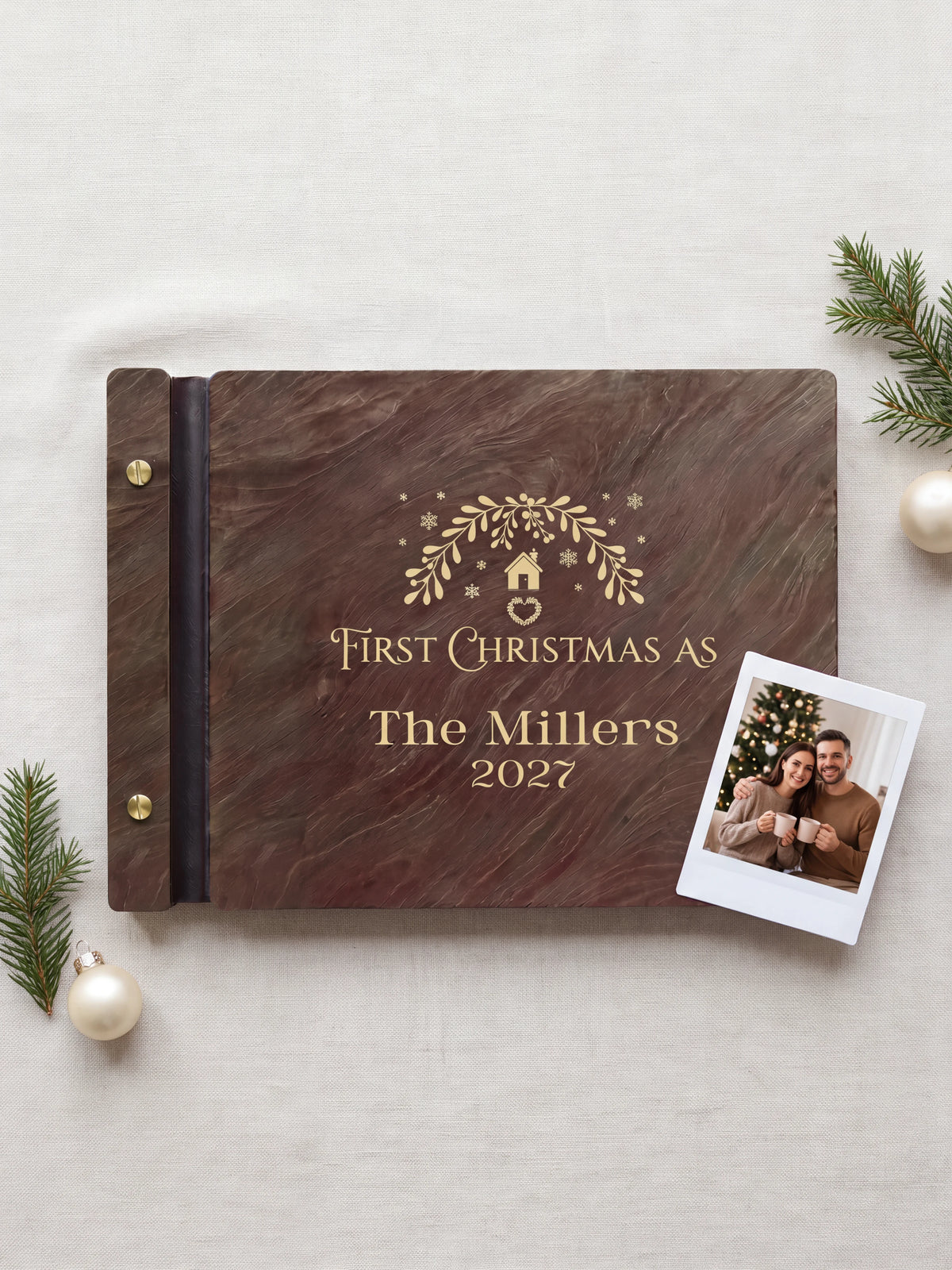 Jacobean wood First Christmas as a married couple photo book lying flat on a coffee table, the cover engraved with “FIRST CHRISTMAS AS”, “The Millers” and “2027”; two gold-tone screws along the black vegan leather spine