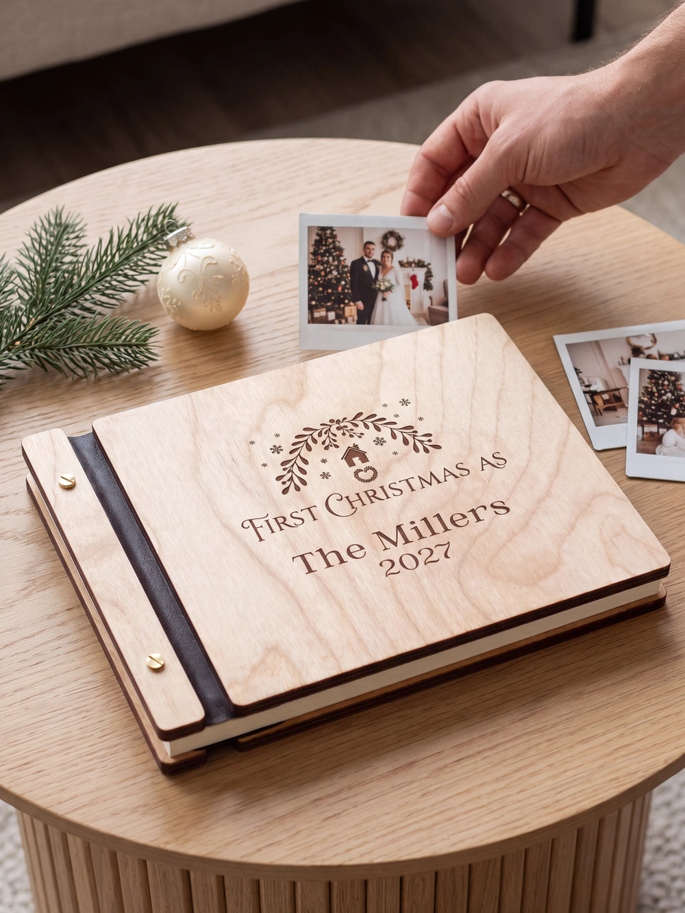 Natural wood First Christmas as a married couple photo book on a light table while a hand picks an instant photo to add; the engraved cover reads “FIRST CHRISTMAS AS”, “The Millers” and “2027”