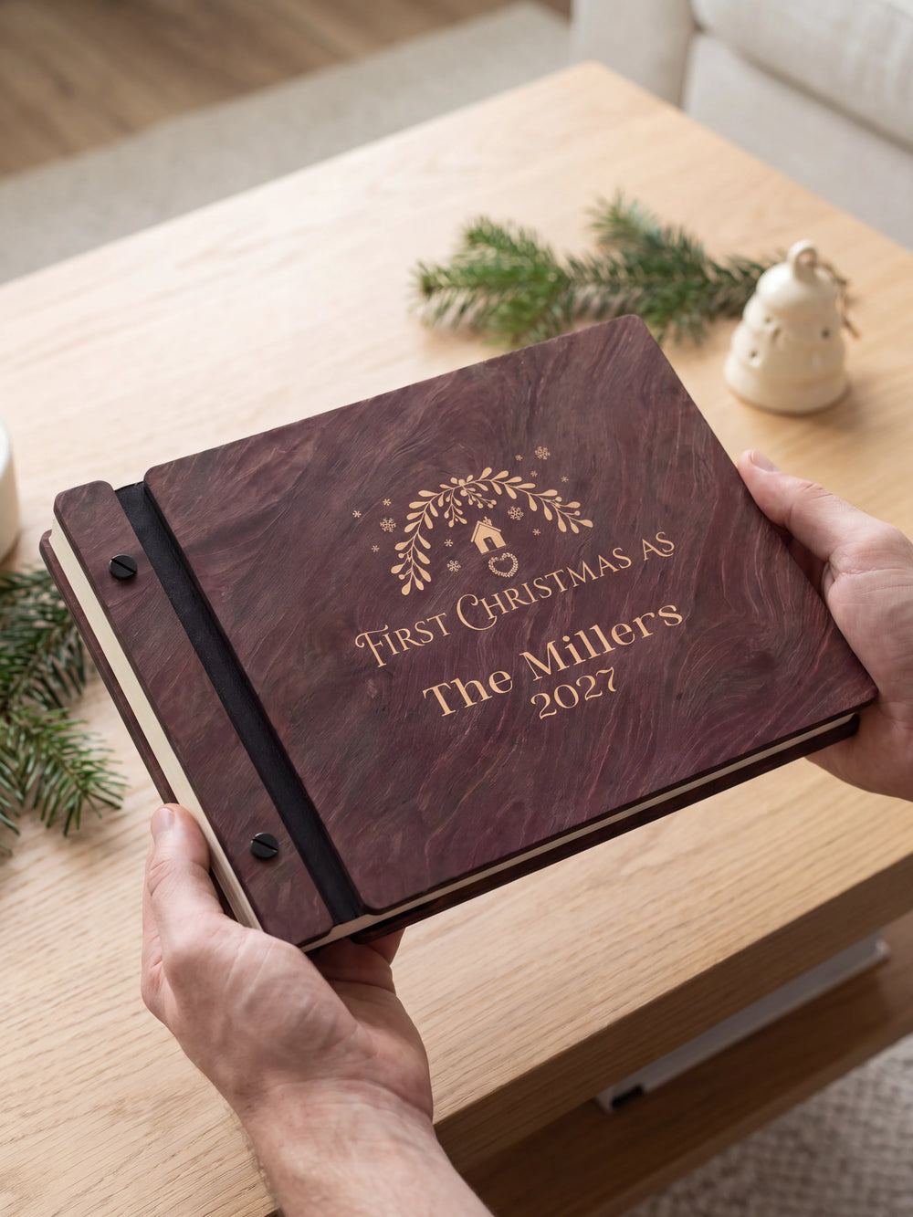 Two hands holding the Jacobean First Christmas as a married couple photo book up to the camera, the cream engraving of a leafy wreath with a tiny house, ornaments and snowflakes and “FIRST CHRISTMAS AS”, “The Millers” and “2027” filling the cover