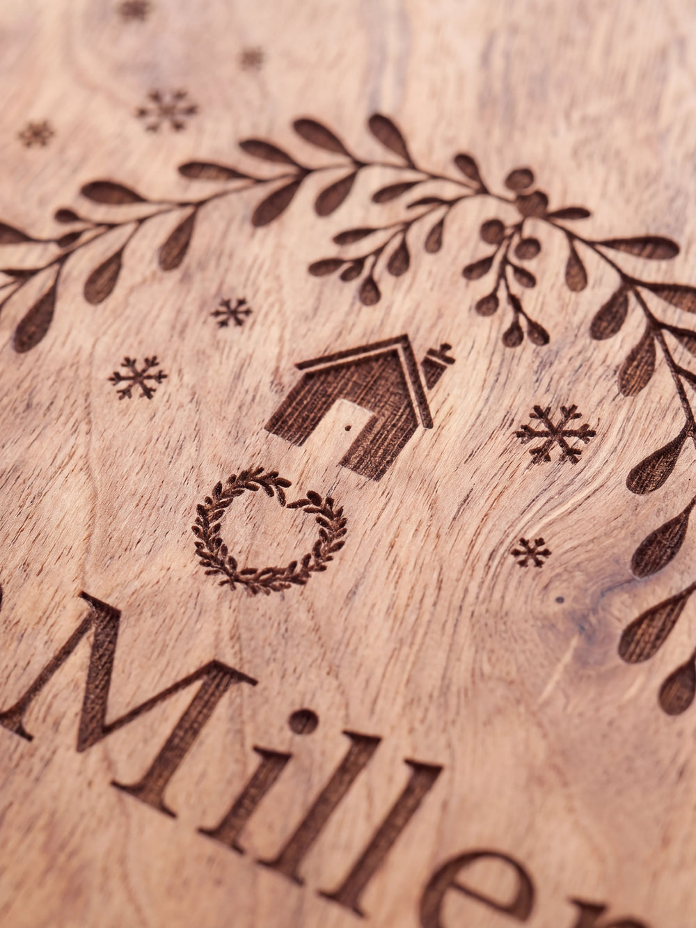 Close-up of the laser engraving on Hazelnut wood: a leafy wreath with a tiny house, ornaments and snowflakes and the personalized text against the visible wood grain