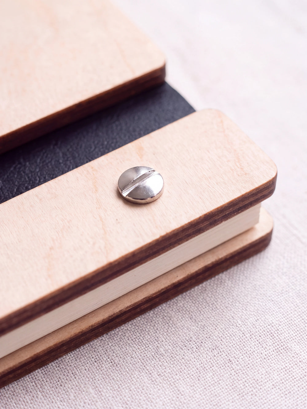 Close-up of a polished silver screw in the Natural wood strip beside the black vegan leather binding, the cream sheet stack visible below
