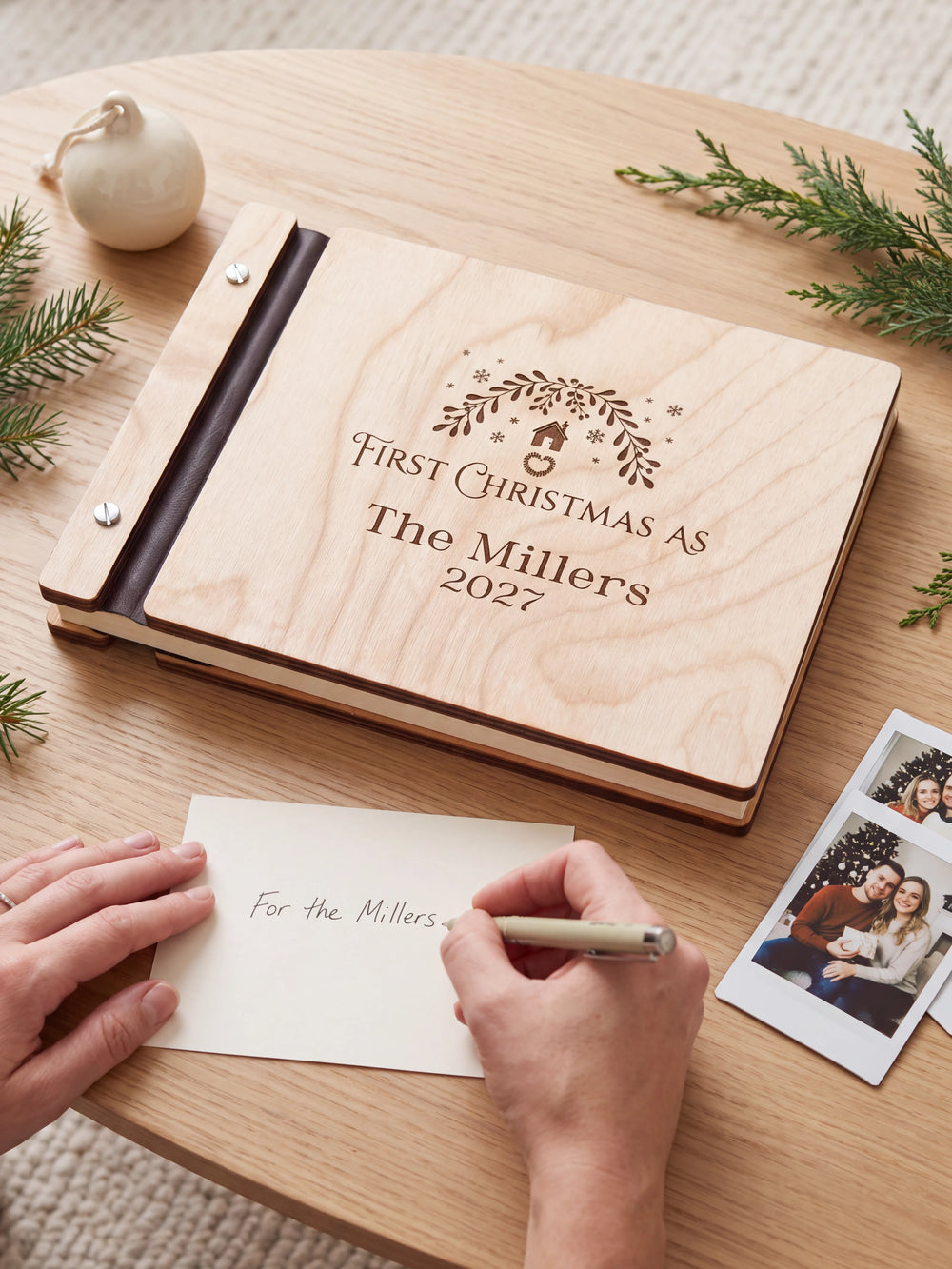A hand writing a small gift card beside the closed Natural wood First Christmas as a married couple photo book; the engraved cover and silver screws are visible