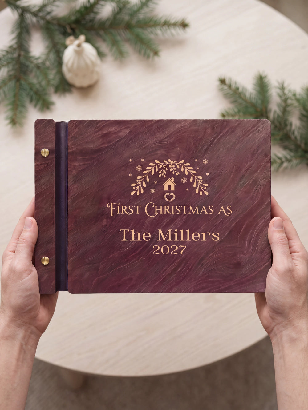 Two hands presenting the closed Jacobean First Christmas as a married couple photo book with its cream engraving of a leafy wreath with a tiny house, ornaments and snowflakes, understated evergreen and an ivory ornament beside them