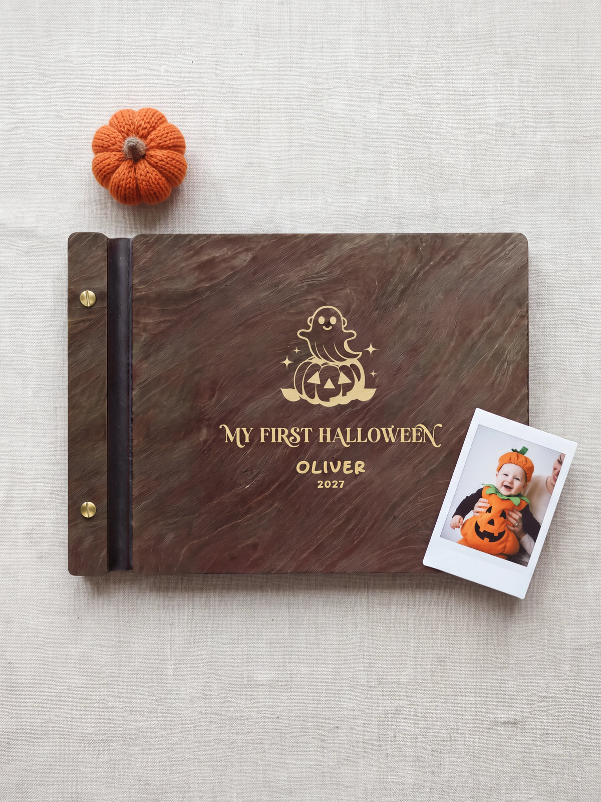 Jacobean wood My First Halloween keepsake book lying flat on oatmeal fabric, the cover engraved with “MY FIRST HALLOWEEN”, “OLIVER” and “2027”; two gold-tone screws along the black vegan leather spine