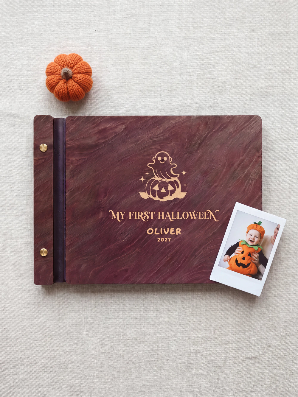 Jacobean wood My First Halloween keepsake book lying flat on oatmeal fabric, the cover engraved with “MY FIRST HALLOWEEN”, “OLIVER” and “2027”; two gold-tone screws along the black vegan leather spine