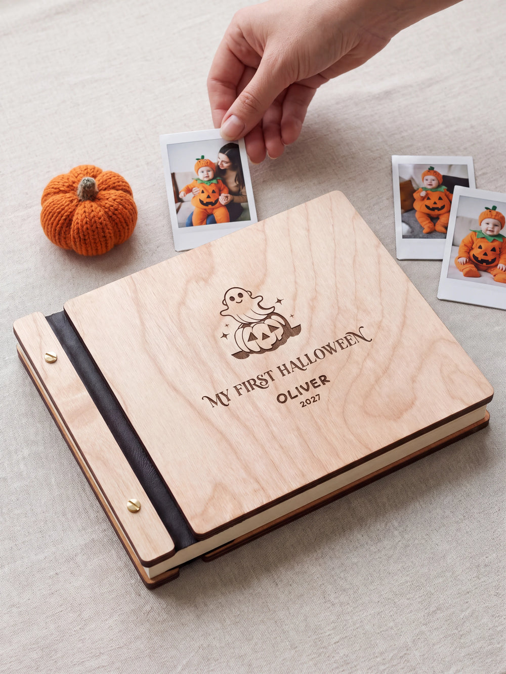 Natural wood My First Halloween keepsake book on a light table while a hand picks an instant photo to add; the engraved cover reads “MY FIRST HALLOWEEN”, “OLIVER” and “2027”