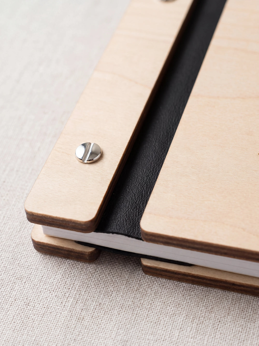 Close-up of a polished silver screw in the Natural wood strip beside the black vegan leather binding, the cream sheet stack visible below