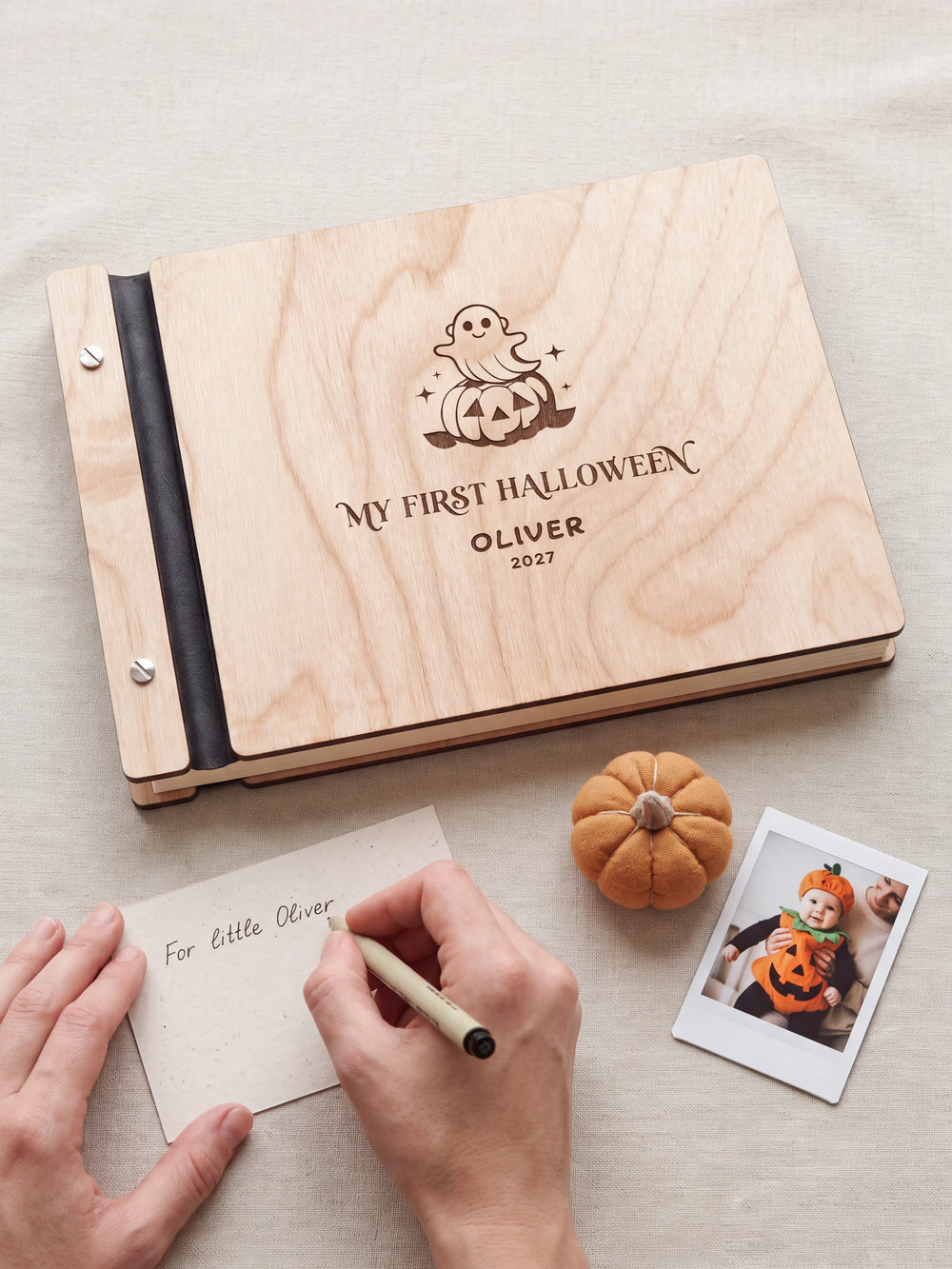 A hand writing a small gift card beside the closed Natural wood My First Halloween keepsake book; the engraved cover and silver screws are visible