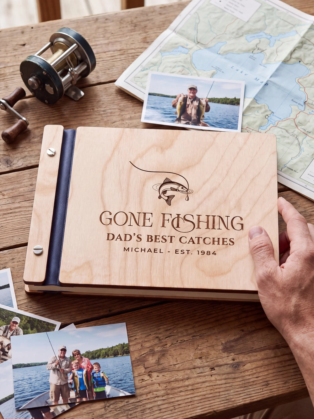 Natural wooden album with brown laser engraving of hooked fish and curved fishing line, shown in a close lifestyle view with navy blue binding and silver screws. The cover reads GONE FISHING.