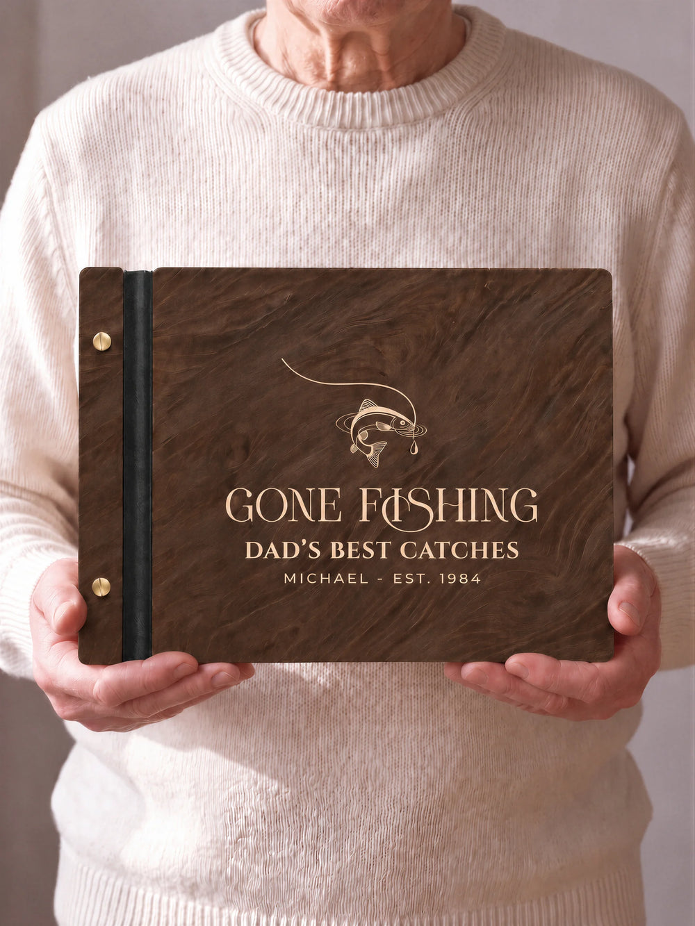 Two hands hold a Jacobean wooden photo album with the GONE FISHING cover facing the camera. Cream engraved artwork, the narrow binding and gold screws are visible.