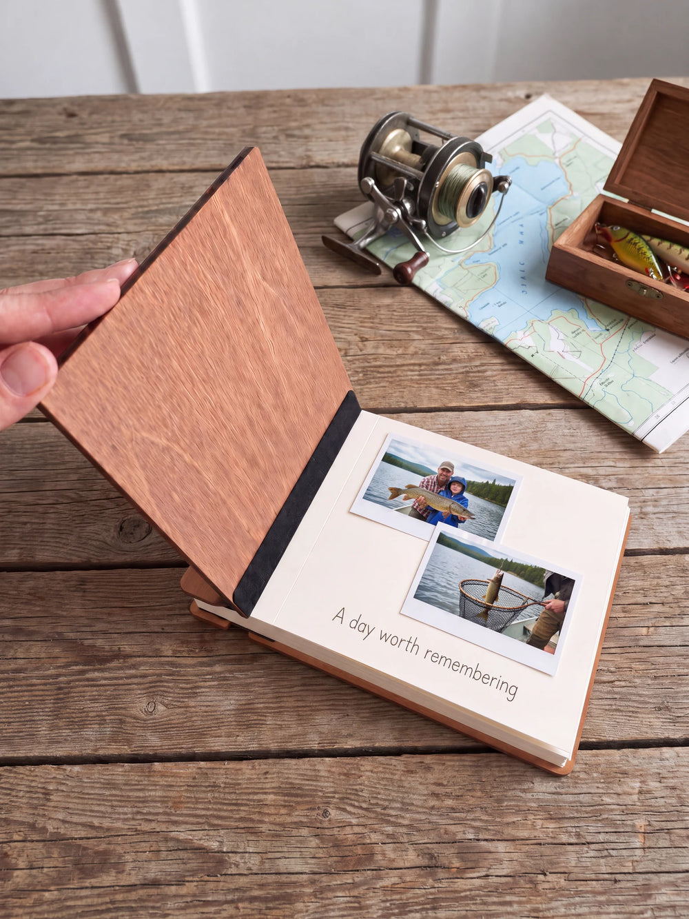 A hand lifts the Hazelnut album’s front cover, revealing a flat black inner fabric strip and a cream page arranged with instant photographs and a handwritten memory.