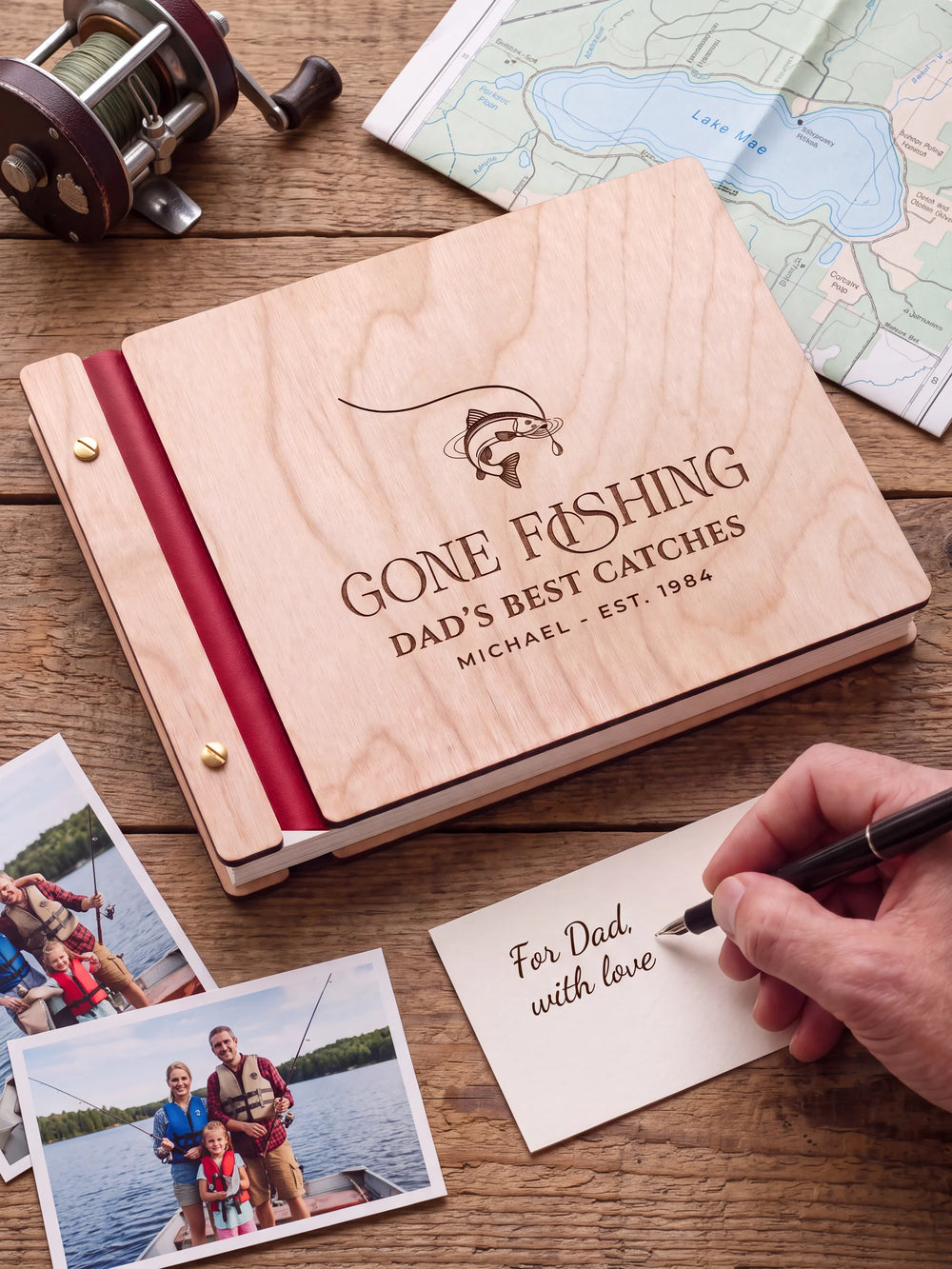 A Natural wooden album with the GONE FISHING engraving, cherry red binding and gold screws. A hand writes on a separate gift card beside the book.