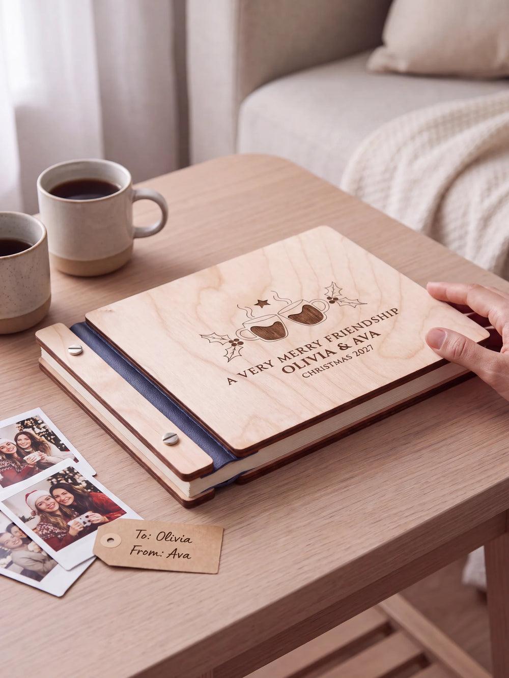 Natural wooden album with brown laser engraving of two steaming mugs with heart centers and holly, shown in a close lifestyle view with navy blue binding and silver screws. The cover reads A VERY MERRY FRIENDSHIP.