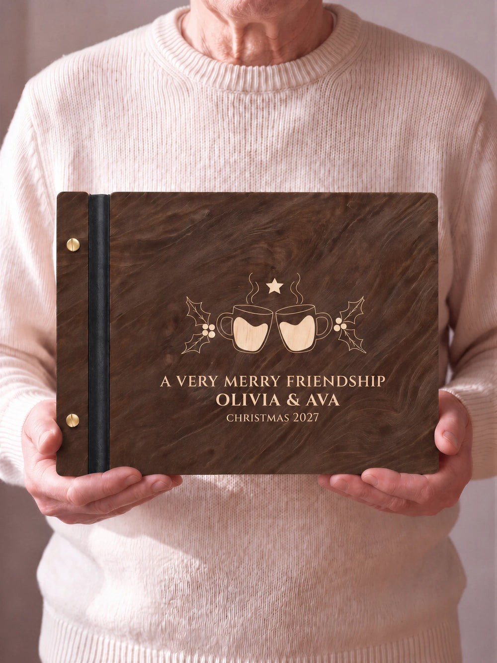 Two hands hold a Jacobean wooden photo album with the A VERY MERRY FRIENDSHIP cover facing the camera. Cream engraved artwork, the narrow binding and gold screws are visible.