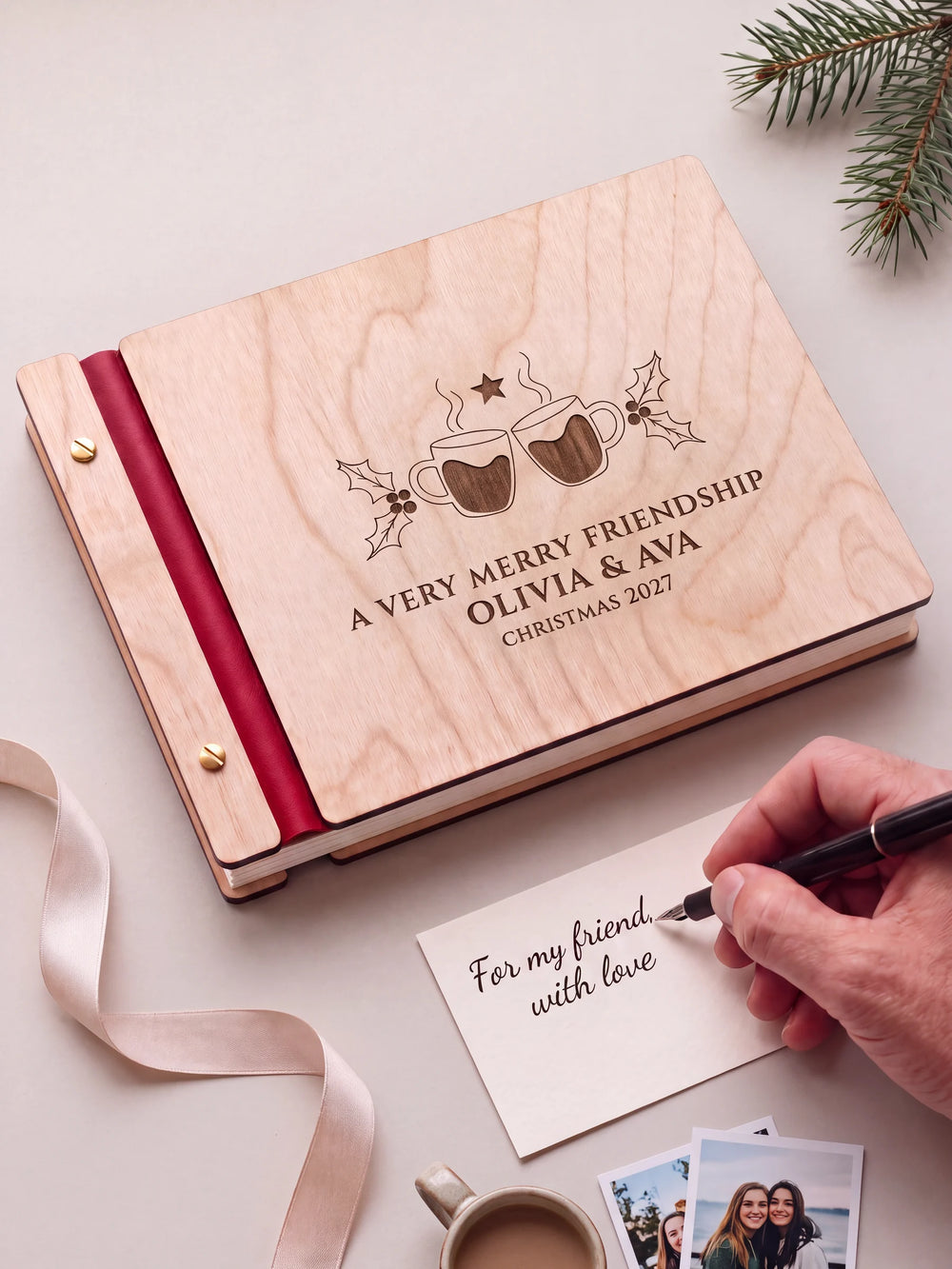 A Natural wooden album with the A VERY MERRY FRIENDSHIP engraving, cherry red binding and gold screws. A hand writes on a separate gift card beside the book.