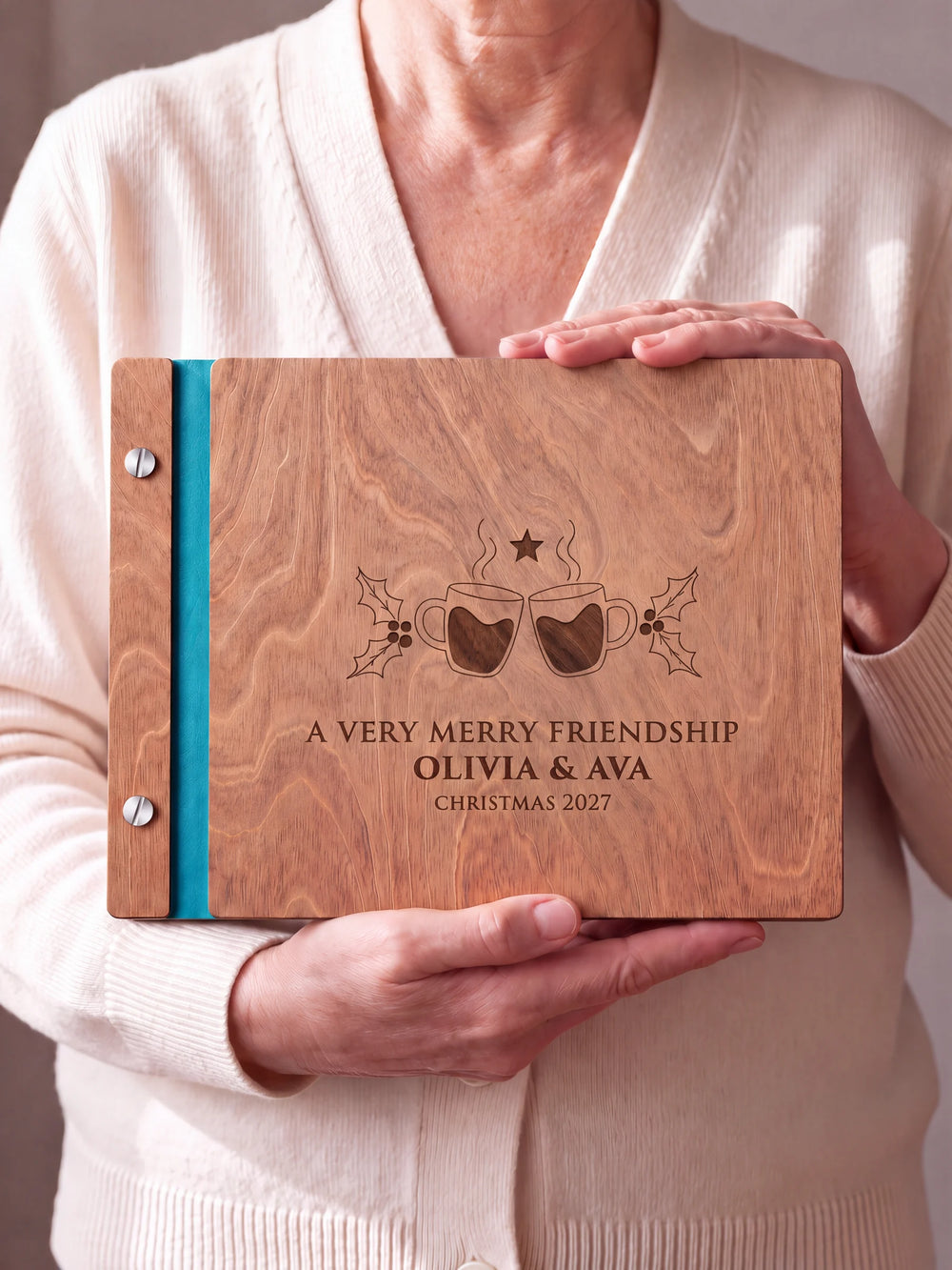 Two hands hold a Hazelnut wooden photo album. The A VERY MERRY FRIENDSHIP cover design has brown engraving, turquoise binding and silver screws.