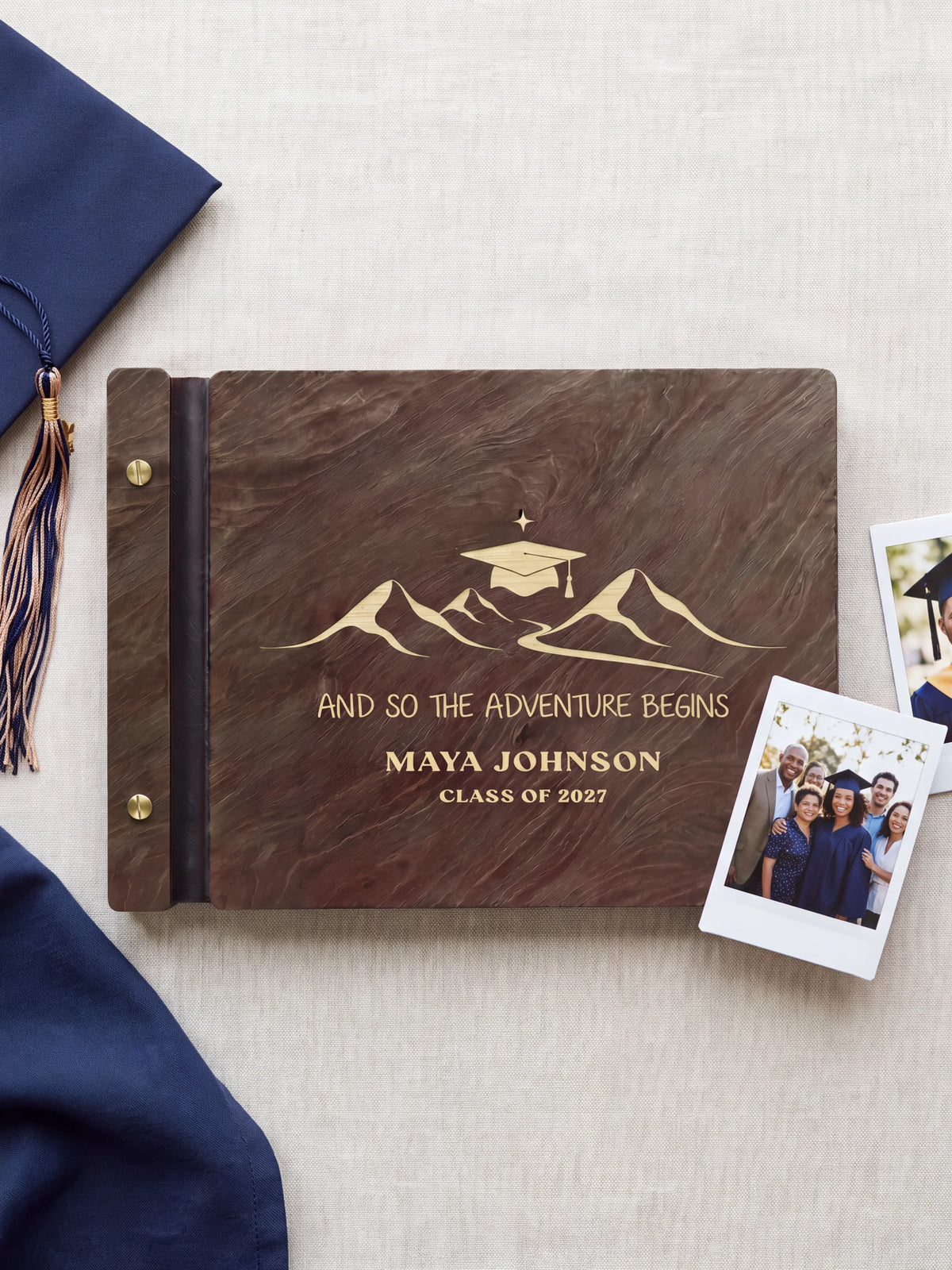 Jacobean wood graduation photo guest book lying flat on navy fabric, the cover engraved with “AND SO THE ADVENTURE BEGINS”, “MAYA JOHNSON” and “CLASS OF 2027”; two gold-tone screws along the black vegan leather spine