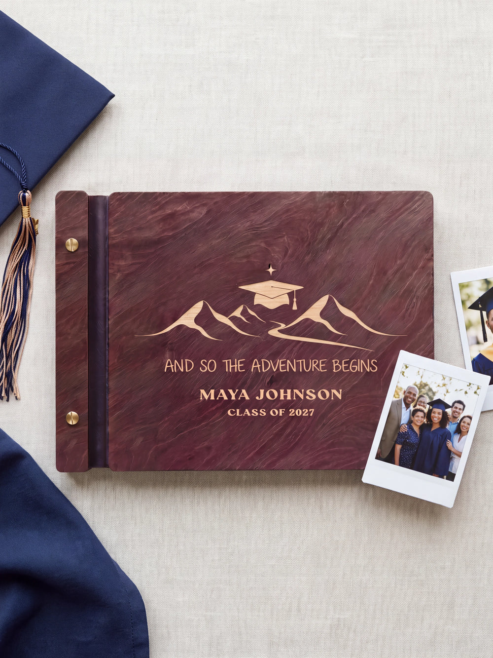 Jacobean wood graduation photo guest book lying flat on navy fabric, the cover engraved with “AND SO THE ADVENTURE BEGINS”, “MAYA JOHNSON” and “CLASS OF 2027”; two gold-tone screws along the black vegan leather spine