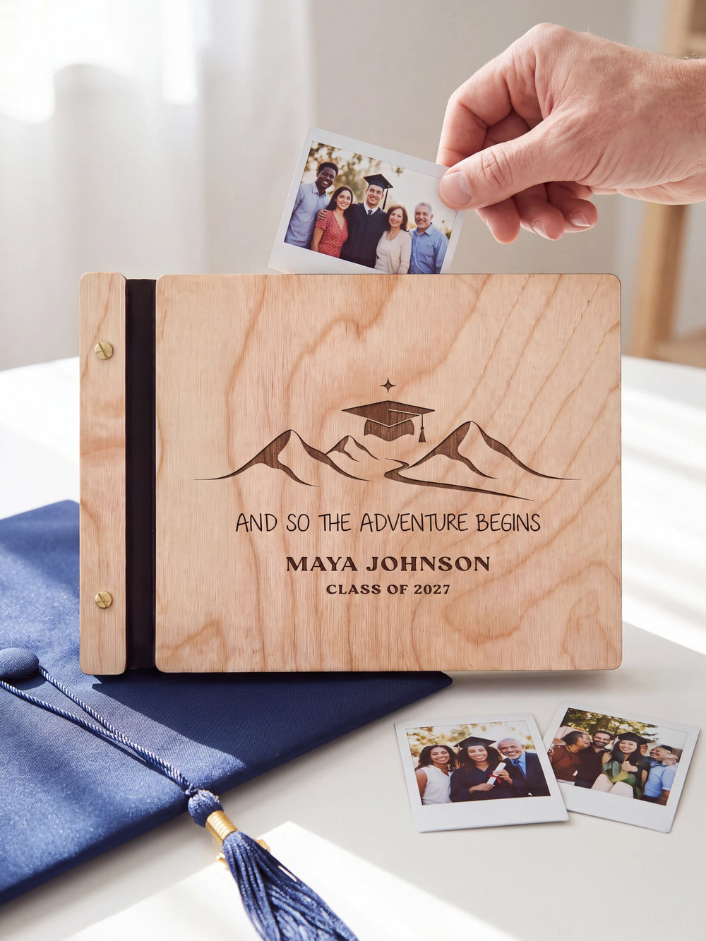 Natural wood graduation photo guest book on a light table while a hand picks an instant photo to add; the engraved cover reads “AND SO THE ADVENTURE BEGINS”, “MAYA JOHNSON” and “CLASS OF 2027”