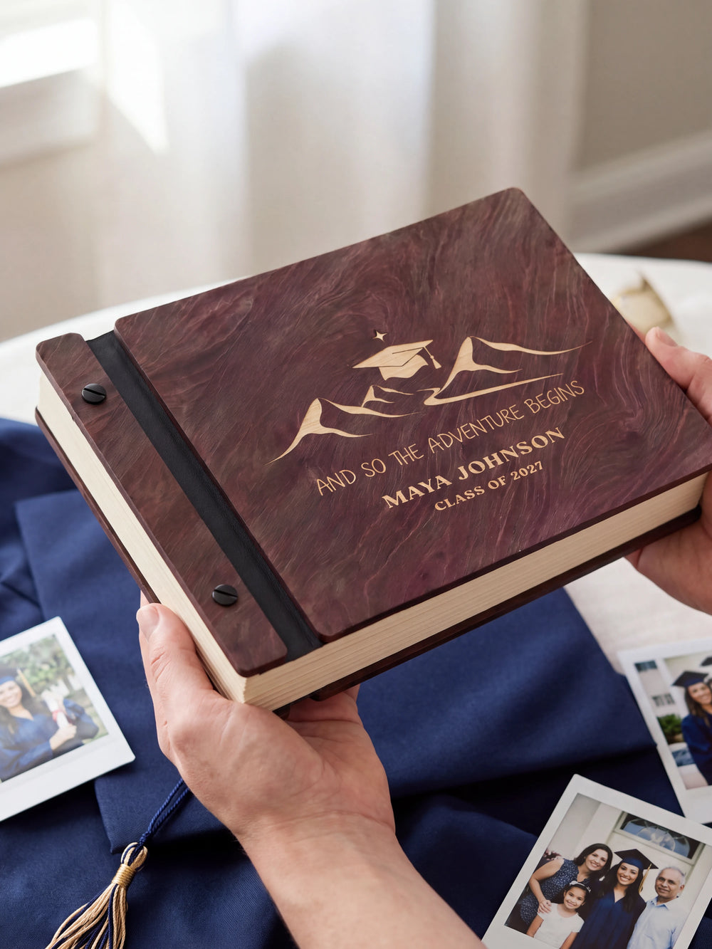Two hands holding the Jacobean graduation photo guest book up to the camera, the cream engraving of mountain peaks beneath a graduation cap and “AND SO THE ADVENTURE BEGINS”, “MAYA JOHNSON” and “CLASS OF 2027” filling the cover
