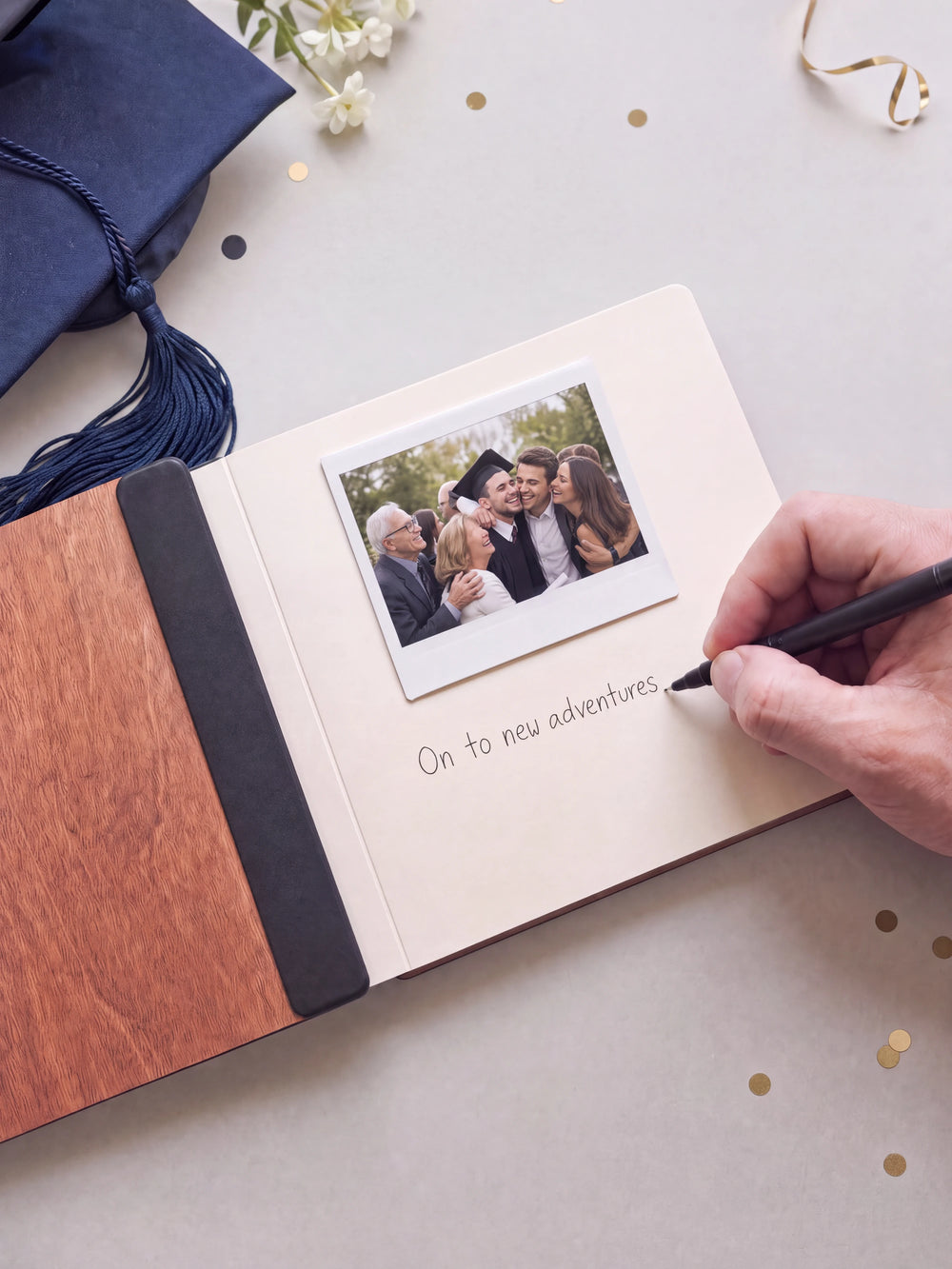 A hand writing a note in fine black pen beneath an instant photo on the cream page of the open Hazelnut graduation photo guest book